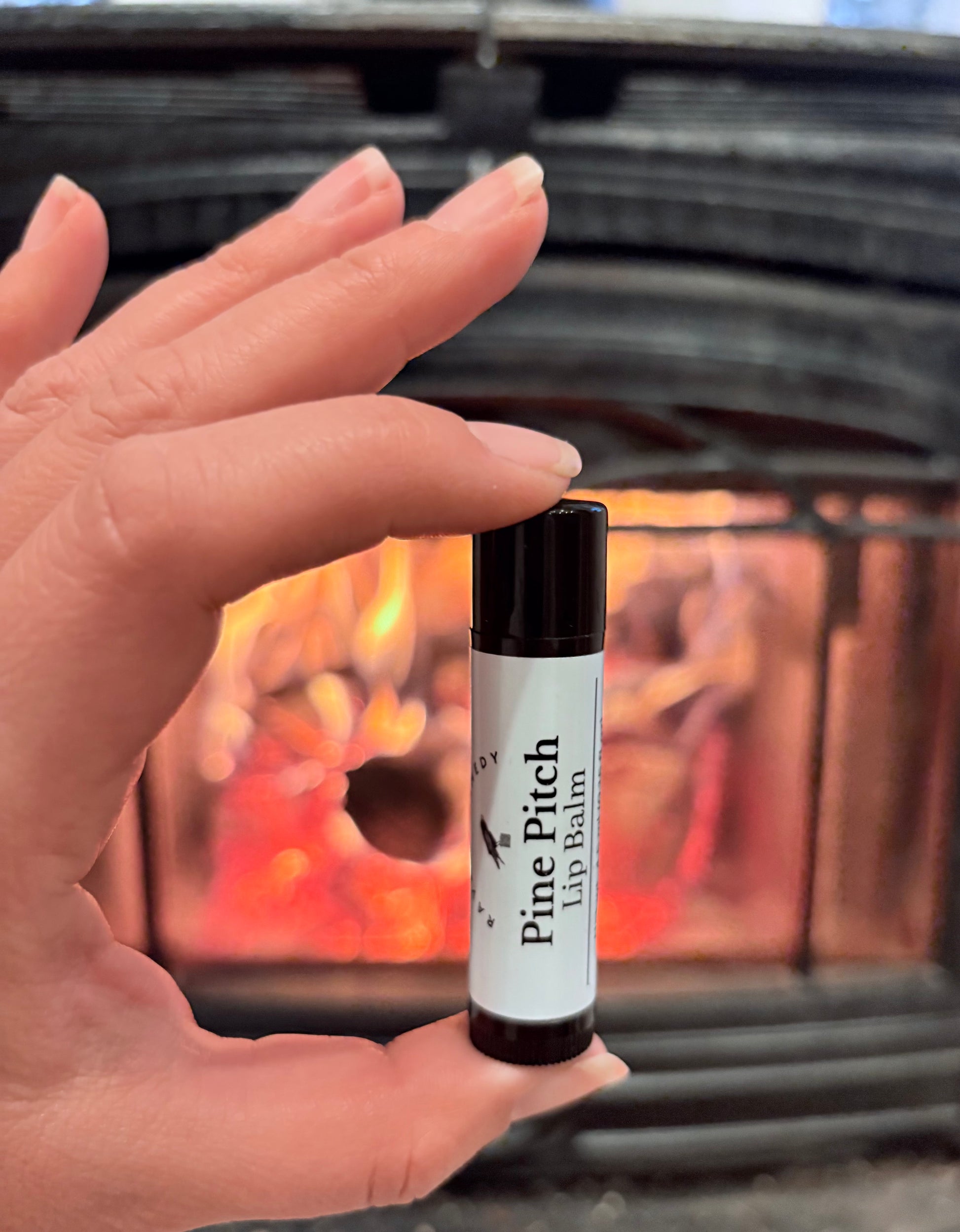 Hand holding a lip balm labeled 'Pine Pitch' in front of a fireplace.