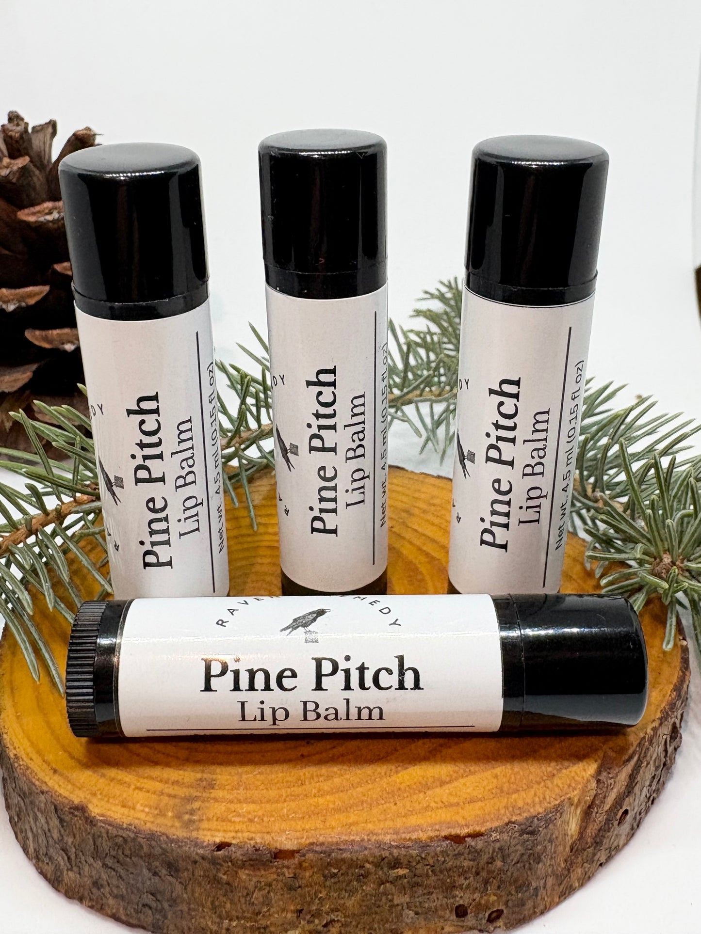 Wildcrafted Pine Pitch Lip Balm tubes displayed on a wood slice with pine sprigs, made by Raven's Remedy.
